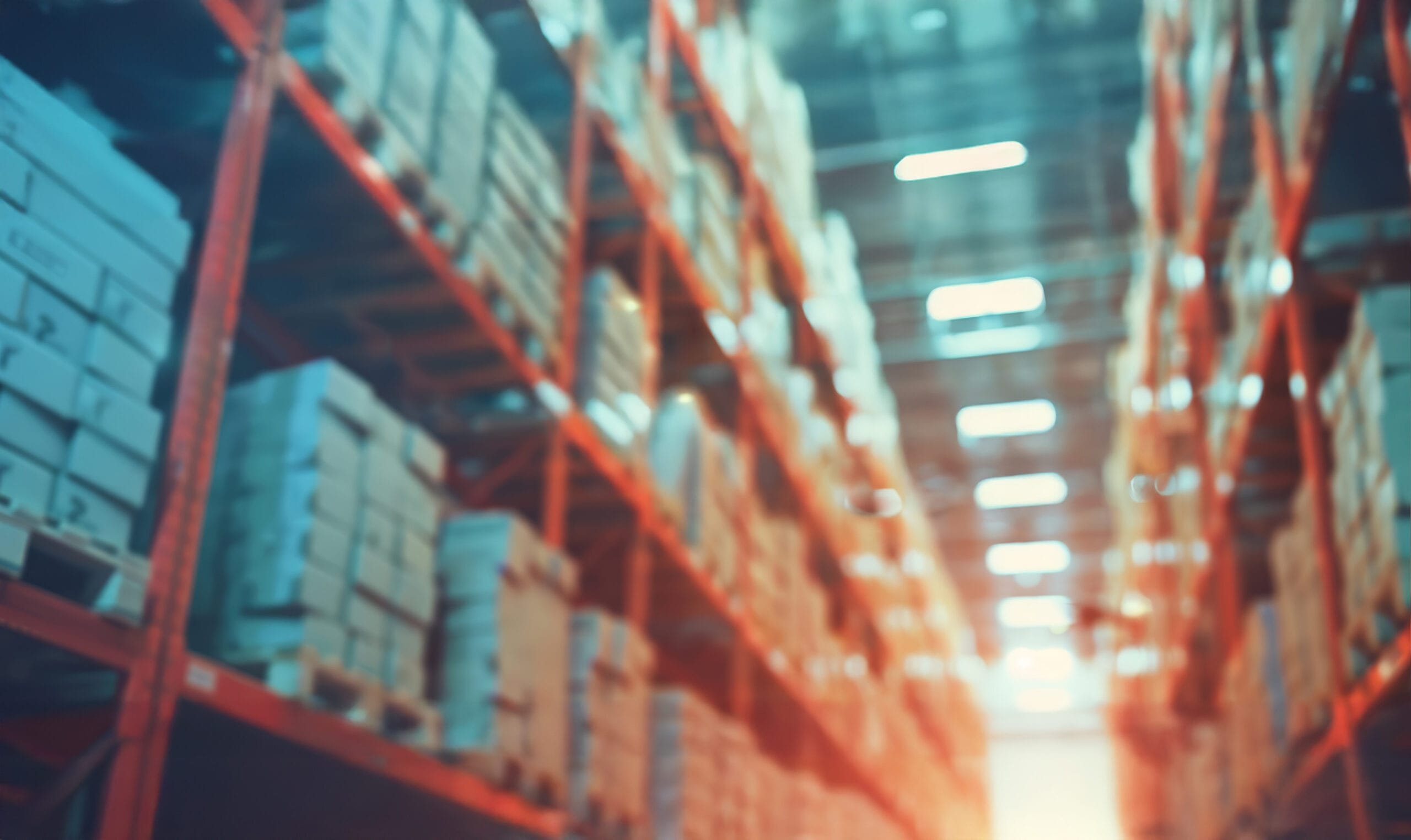 Aisle in a warehouse distribution center lined with tall storage racks stacked with boxes, lit by rows of overhead lighting